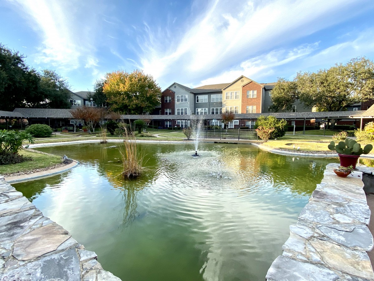 A serene pond with a fountain and surrounding buildings under a blue sky.
