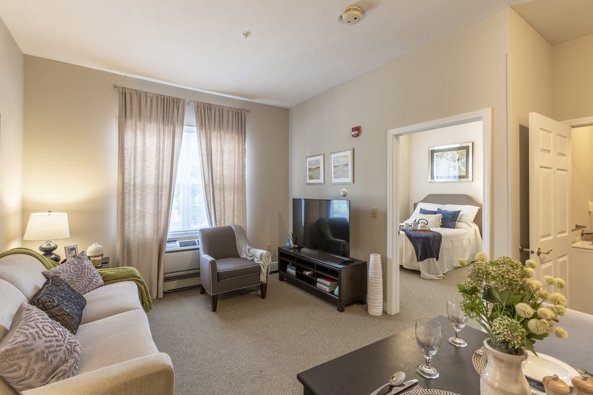 A cozy living room with modern decor, featuring a comfortable armchair, neutral-colored curtains, and a large television. The entrance to a bedroom is visible in the background.