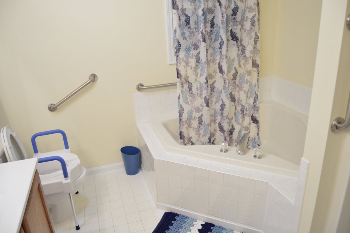 An accessible bathroom featuring a bathtub with grab bars, a shower curtain, a safety chair, and a small trash bin on the floor.