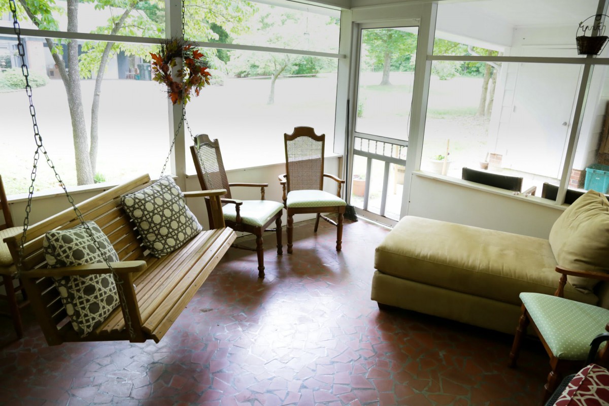 A cozy porch with a wooden swing, chairs, and an ottoman bathed in natural light.