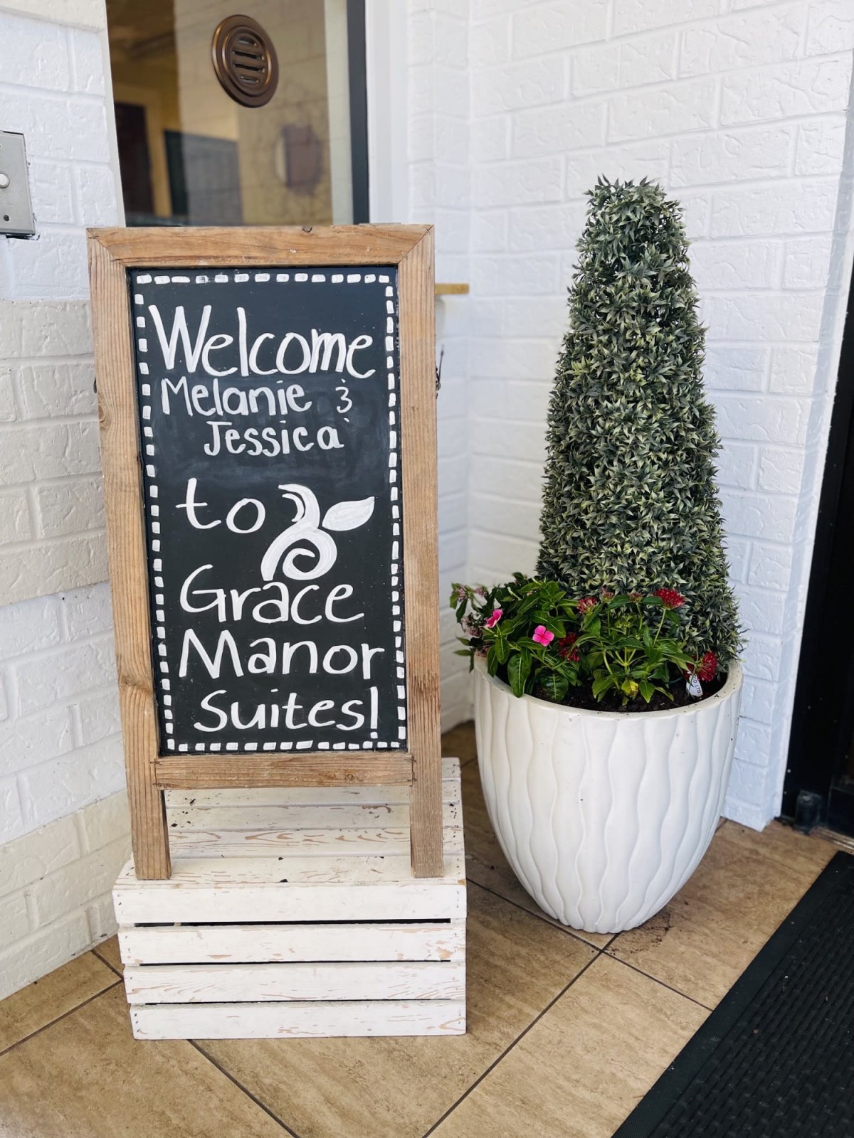 A welcoming chalkboard sign at Grace Manor Suites with a potted plant beside it.
