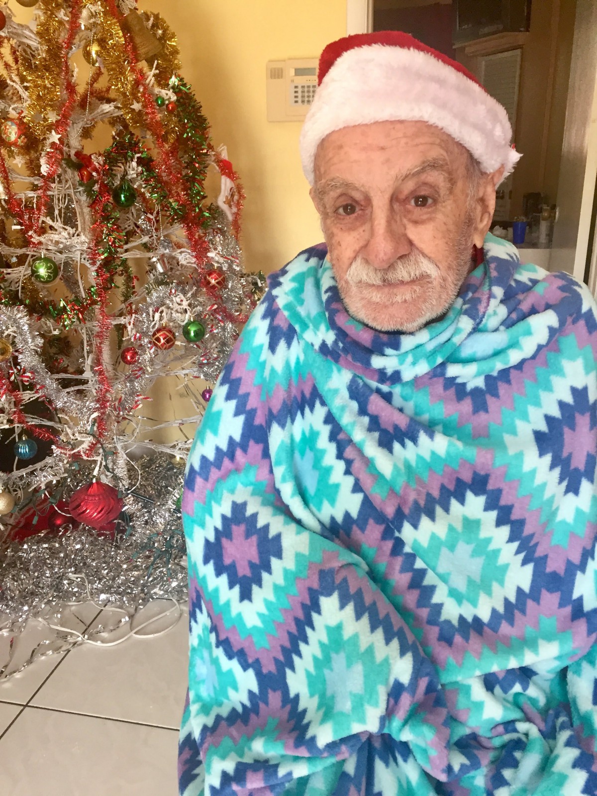 An elderly person wrapped in a colorful blanket sits near a decorated Christmas tree, creating a festive atmosphere indoors.