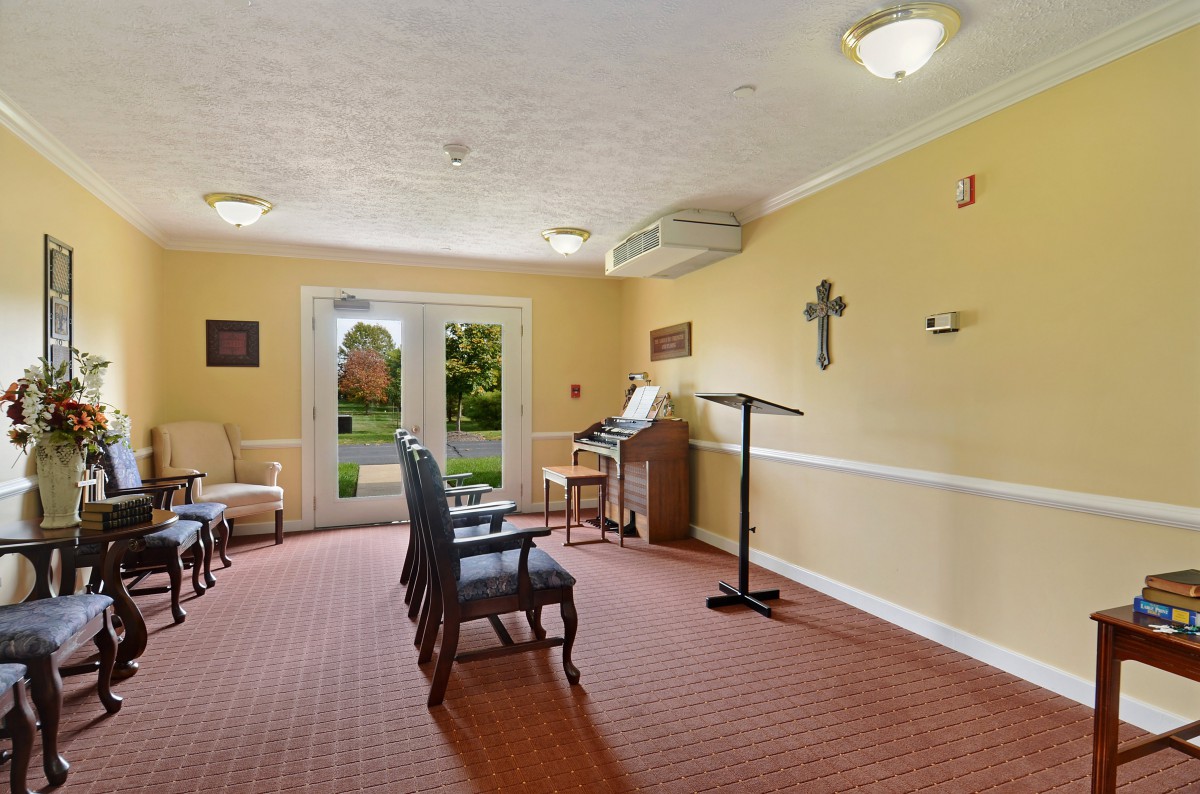 A small chapel interior with chairs, a cross on the wall, and an organ near the window.