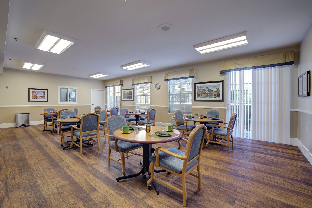A well-lit dining room with multiple tables and chairs arranged neatly, featuring large windows and wooden flooring.