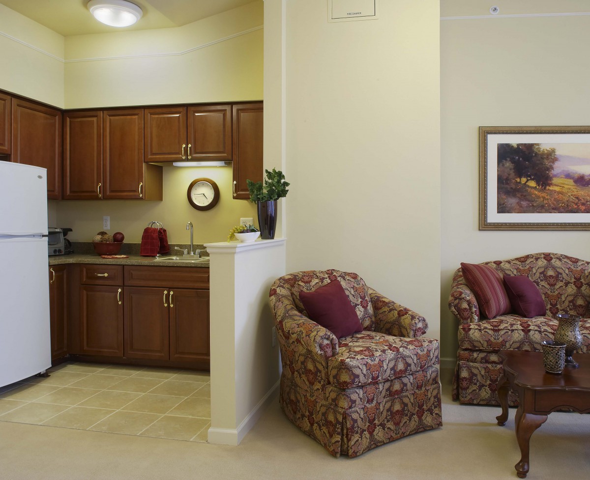A cozy living room leading to a small kitchen area, featuring comfortable armchairs and wooden cabinets.