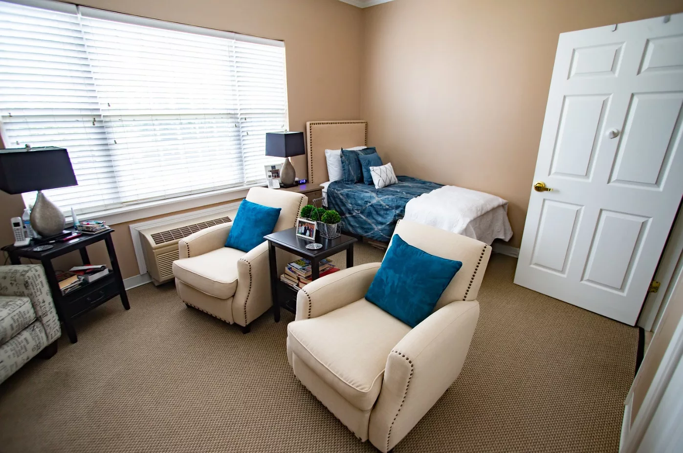 A cozy bedroom with a neatly made bed and two comfortable chairs, complemented by soft lighting from a large window.