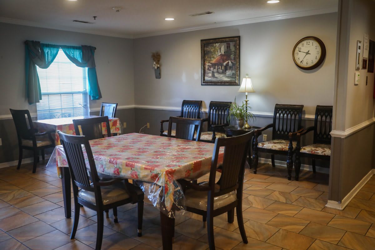 A cozy dining area with a table covered in a patterned cloth surrounded by chairs, featuring a wall clock, painting, and a lamp for decoration.