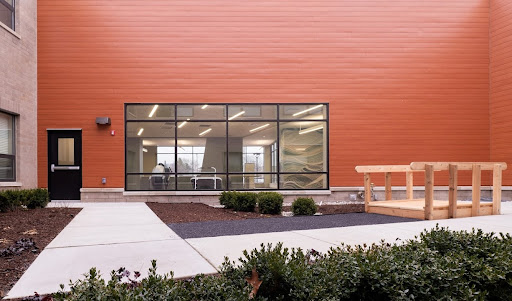 A modern building featuring large windows and an orange exterior, with well-maintained landscaping in front.