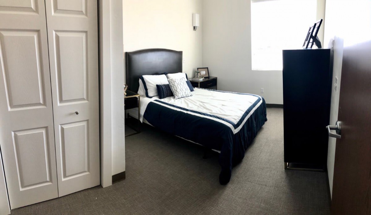A neatly arranged bedroom featuring a bed with dark bedding, nightstands on either side, and natural light coming through a window.
