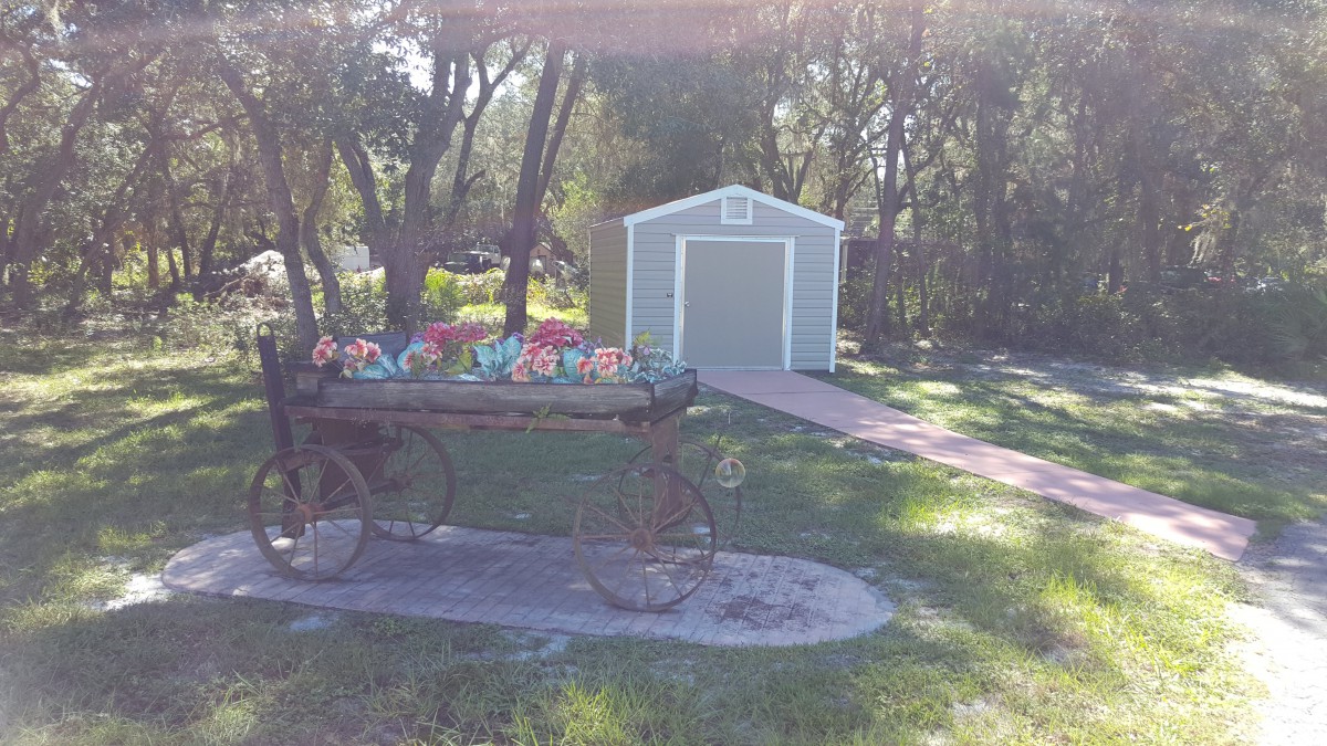 A rustic wagon decorated with flowers stands in a garden, with a shed in the background surrounded by trees.