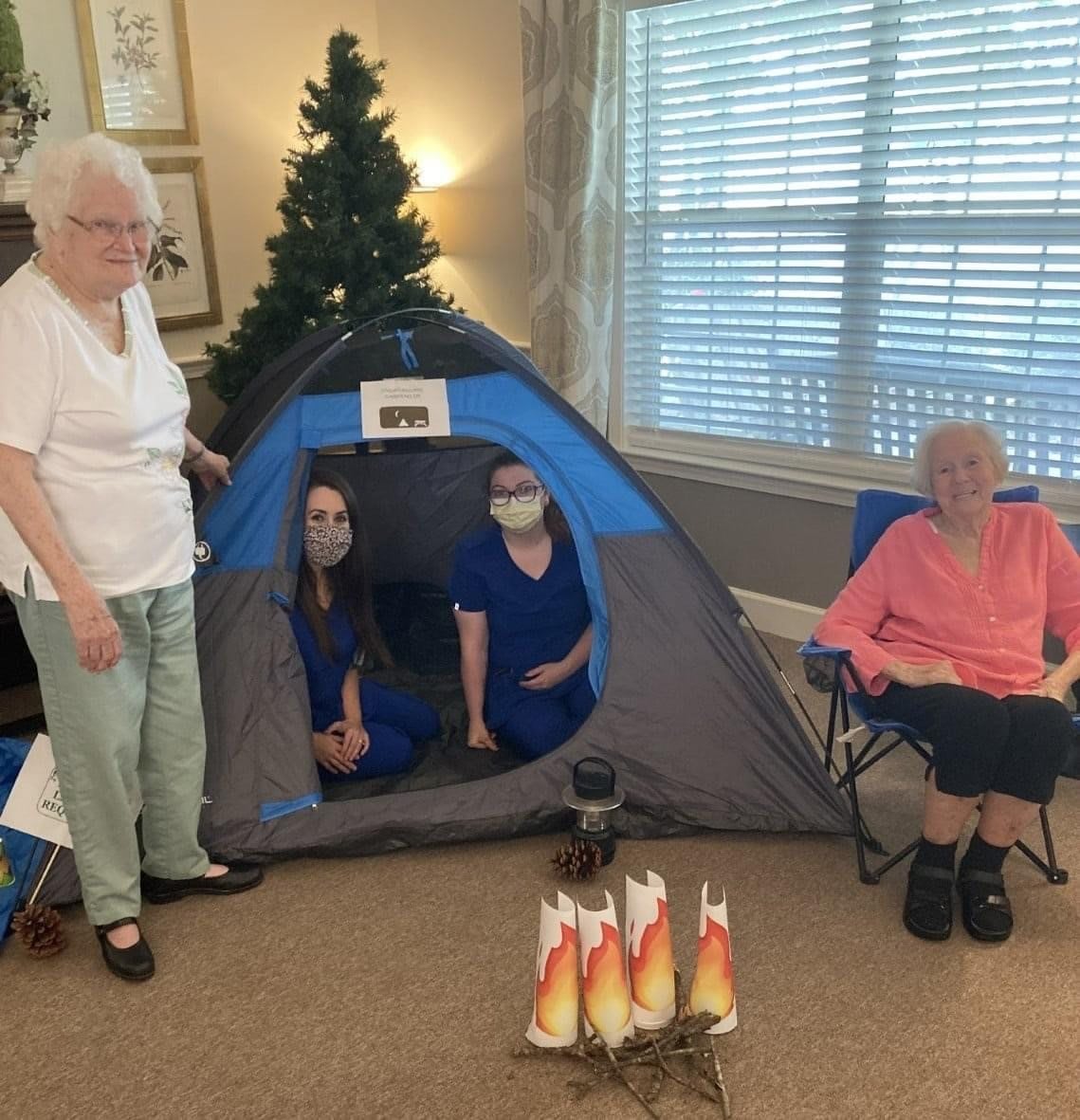 A cozy indoor camping setup with a tent and an artificial campfire, featuring a small group of people in a living room.