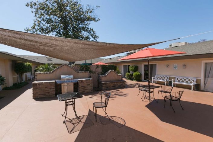 A sunny outdoor patio area with tables and chairs, shaded by a large sail canopy.