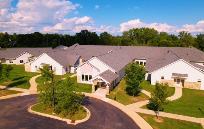 An aerial view of a residential building surrounded by lush greenery and a winding driveway under a partly cloudy sky.
