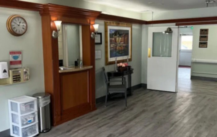 A small reception area featuring a wooden desk, a chair, a painting on the wall, and a door leading to another room.