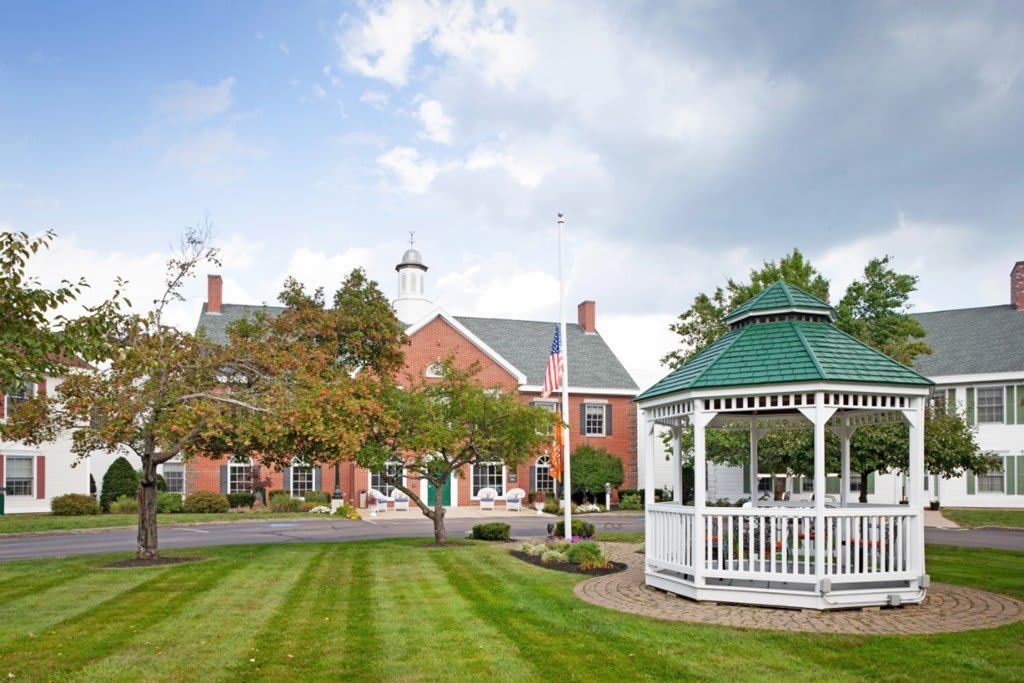 A scenic view of a gazebo on a well-manicured lawn with a brick building and trees in the background under a cloudy sky.