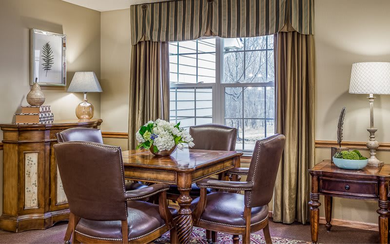 A cozy dining room with a wooden table and leather chairs, adorned with flowers, and illuminated by natural light through the window.
