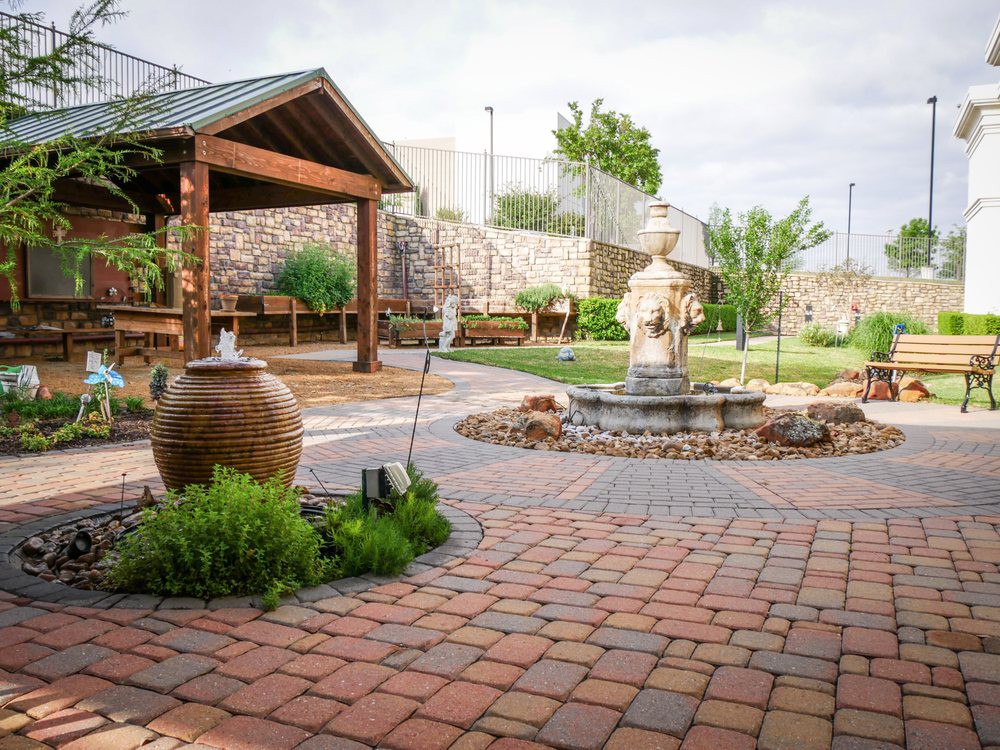 A serene garden courtyard with a central fountain, surrounded by lush greenery and a paved walkway.