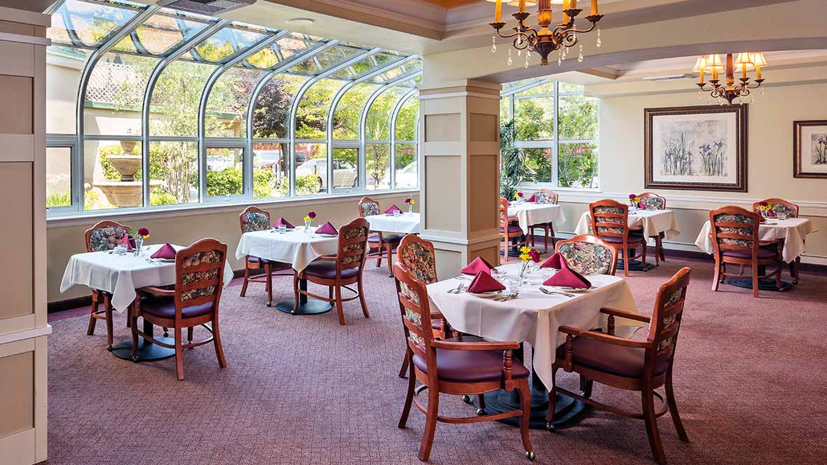 An elegant restaurant dining area with tables set beneath large windows and a chandelier.
