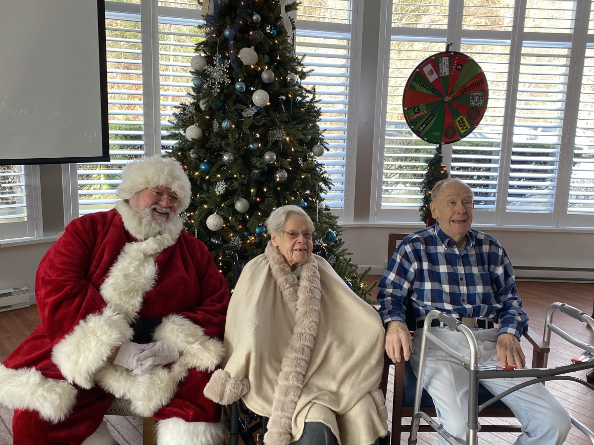 A festive indoor setting with Santa Claus and two elderly individuals sitting by a decorated Christmas tree.