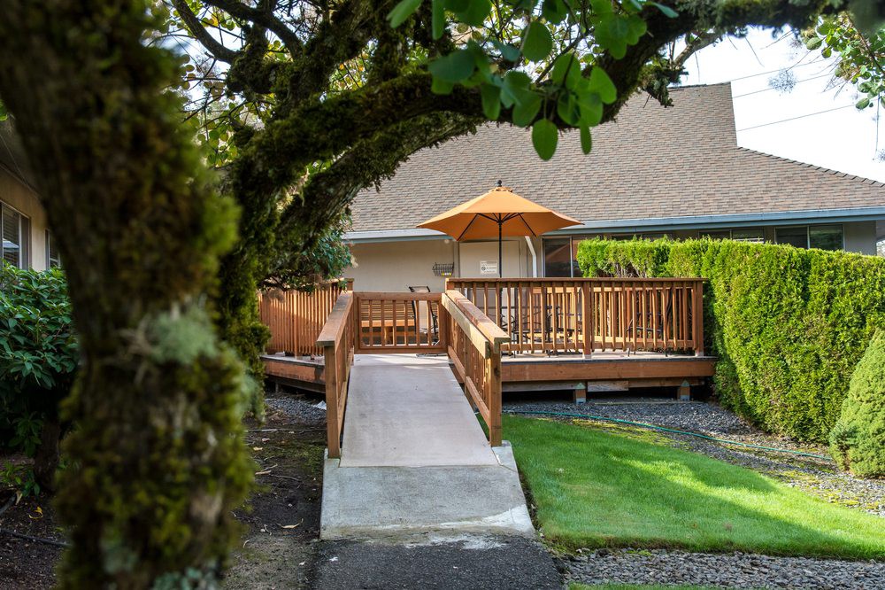 A serene outdoor deck with a ramp, shaded by an umbrella, surrounded by lush greenery.