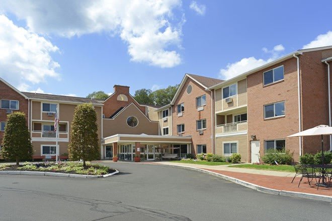 A multi-story residential building with a central entrance and surrounding parking area under a partly cloudy sky.