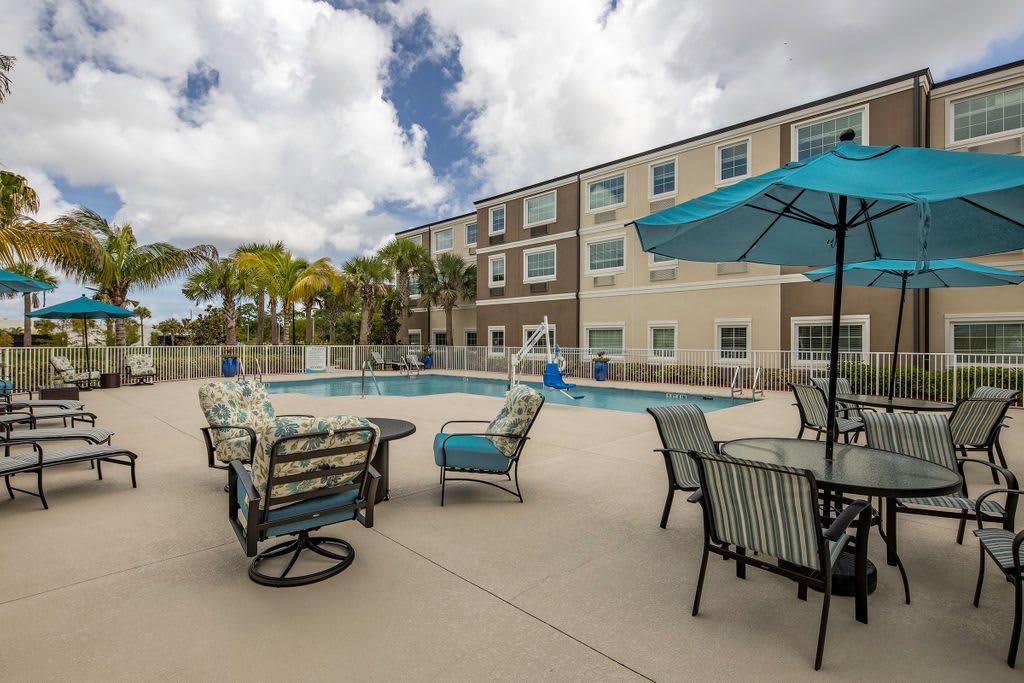 A view of an outdoor swimming pool with a seating area and umbrellas, situated in front of a multi-story apartment building under a cloudy sky.