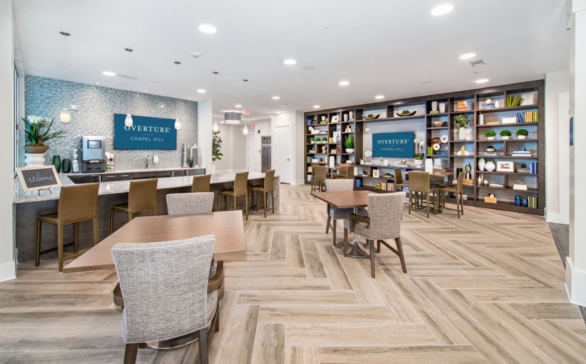 A modern cafe interior with wooden flooring and bookshelves, featuring ample seating and bright lighting.
