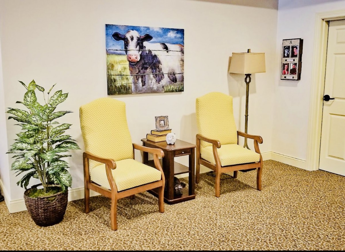 A cozy seating area with two yellow chairs, a small table, a standing lamp, and a cow painting on the wall.