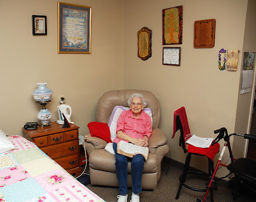 An elderly person sitting comfortably in an armchair, surrounded by personal items and wall decorations.