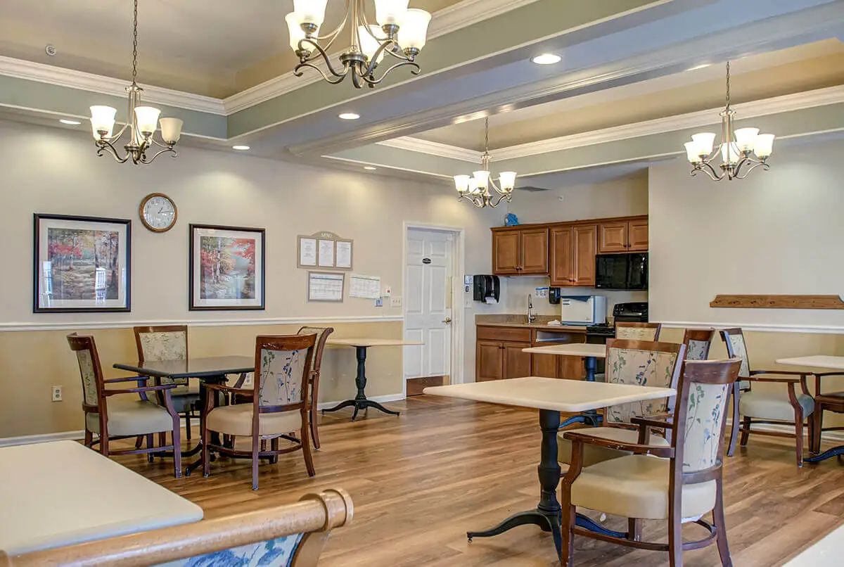 A well-lit dining area with tables and chairs, adjacent to a kitchen with wooden cabinets and chandeliers overhead.