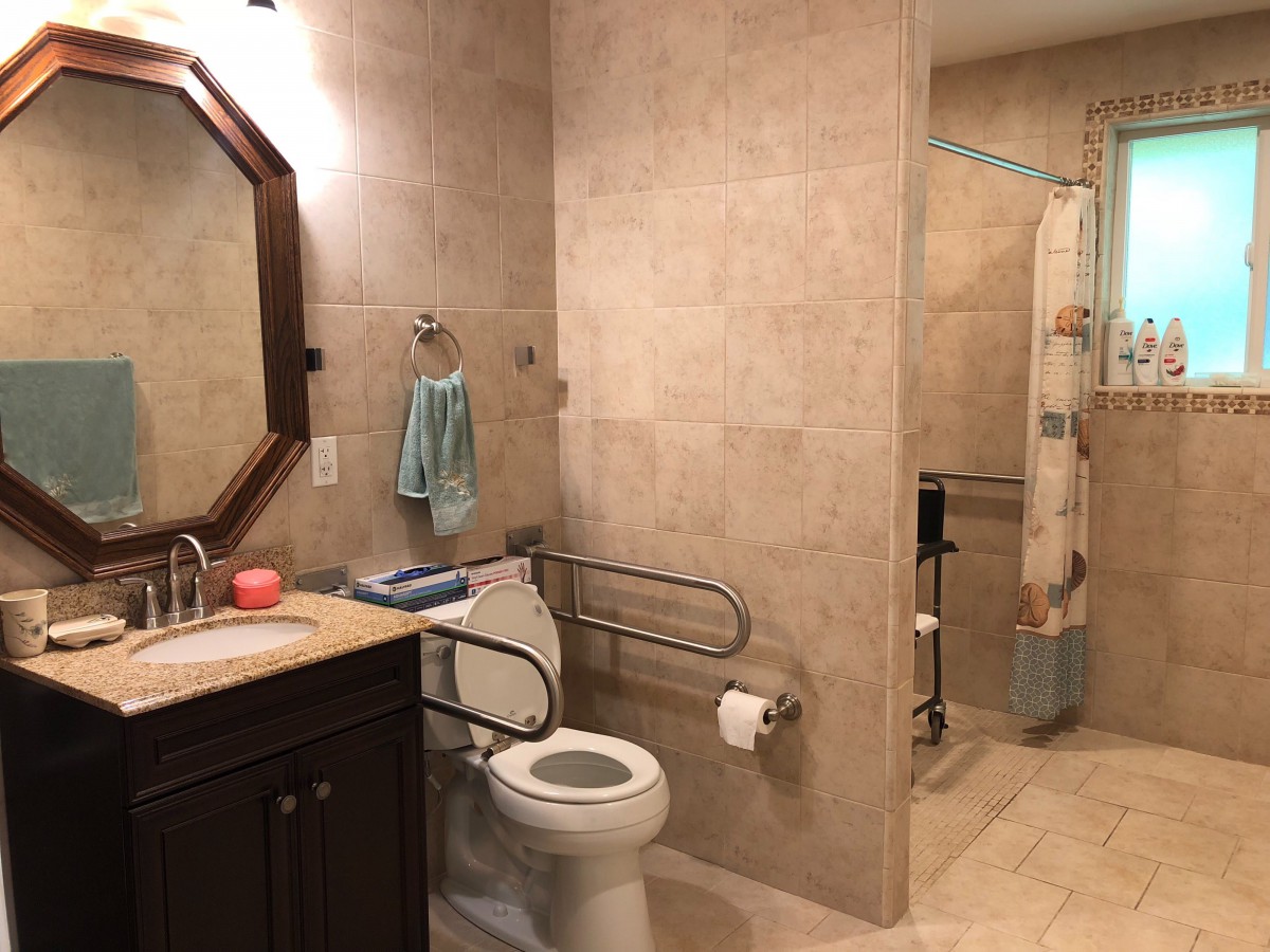An accessible bathroom with tiled walls and flooring, featuring a toilet with handrails, a sink with a mirror above, and a shower area.