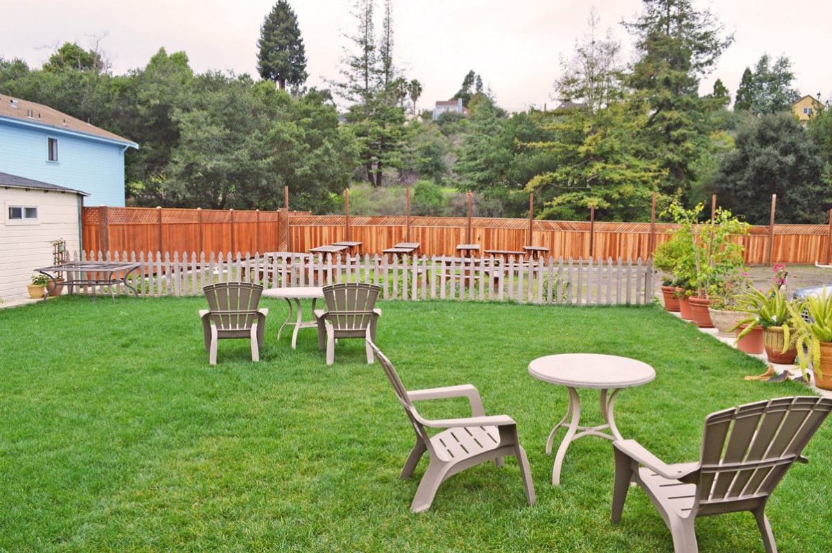 An outdoor garden with plastic chairs and tables set on a lush green lawn, surrounded by a wooden fence and trees.