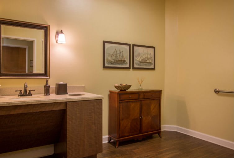 A warmly lit bathroom featuring a wooden cabinet, artwork on the wall, and a countertop with a mirror and light fixture.