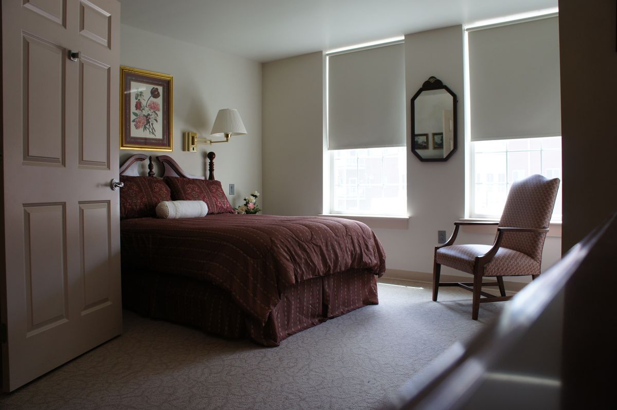 A cozy bedroom with a neatly made bed, chair, and windows with blinds.