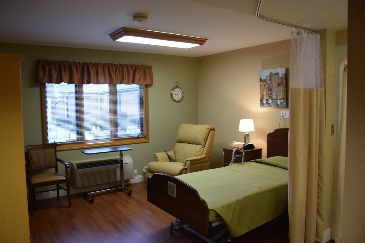 A well-maintained hospital room with a single patient bed, comfortable chair, and natural light from the window.