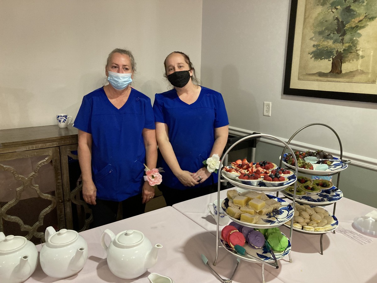Two people wearing blue uniforms and masks stand next to a table set with desserts and a tea set.