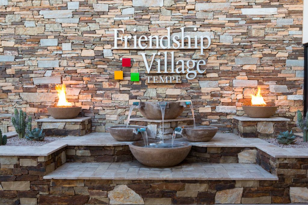 A decorative water fountain with fire bowls in front of a stone wall displaying the name 'Friendship Village Tempe'.
