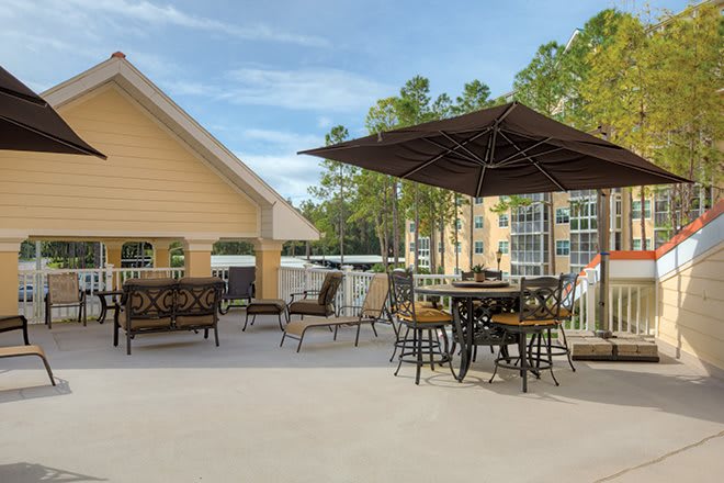A spacious outdoor patio with tables, chairs, and an umbrella, surrounded by trees and adjacent to a building.