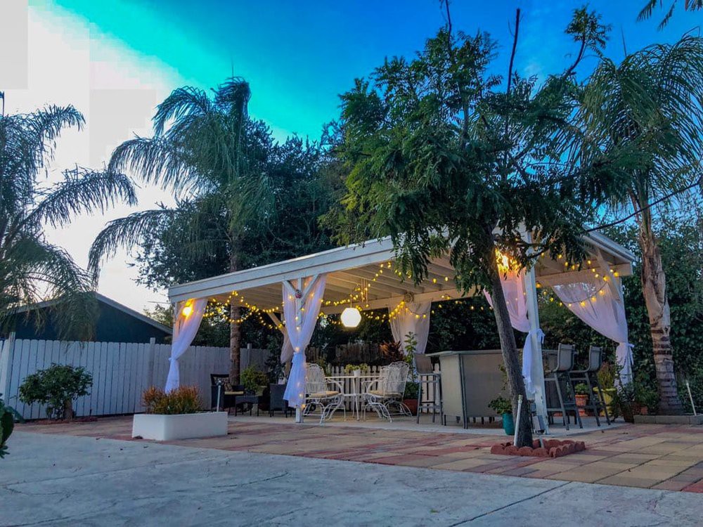 A cozy outdoor patio area with a pergola, draped curtains, and warm lighting surrounded by lush palm trees.
