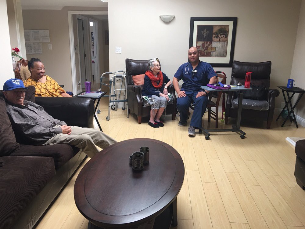 A group of elderly people and a healthcare worker sitting in a nursing home common area.