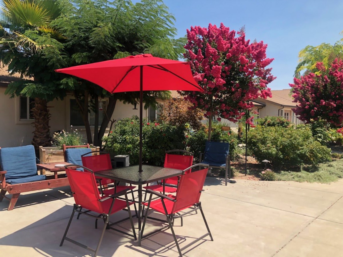 A sunny patio with a red umbrella and matching chairs, surrounded by lush greenery and flowering trees.