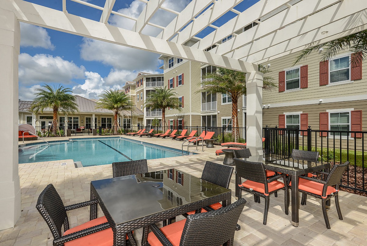 A sunny outdoor pool area with lounge chairs and patio furniture at an apartment complex.