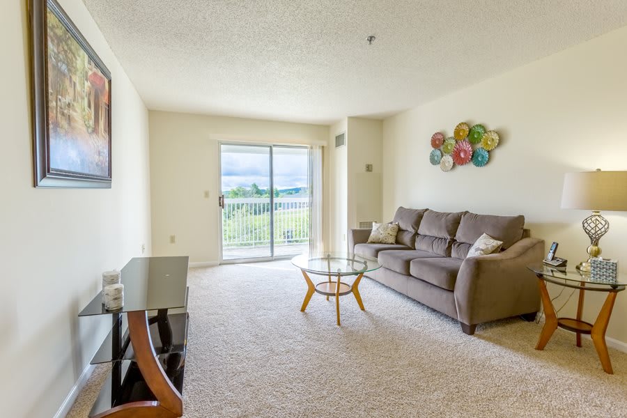 A bright living room featuring a comfortable sofa, a modern coffee table, and colorful artwork on the wall. The space opens to a balcony with a scenic view.