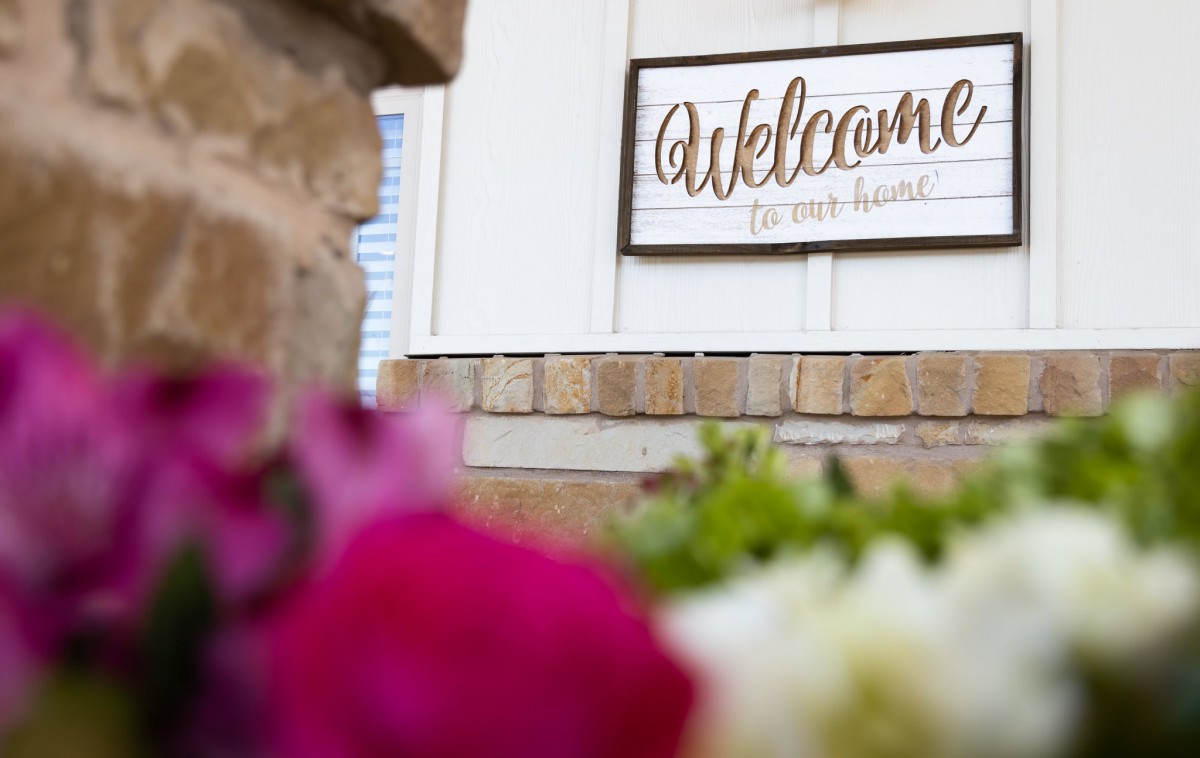 A welcoming sign on a brick wall surrounded by vibrant flowers.