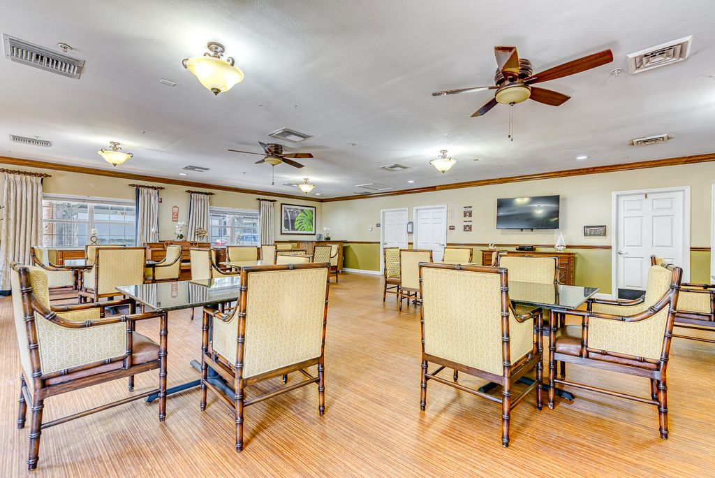 A spacious dining room with multiple tables and chairs, featuring ceiling fans and a wall-mounted television.