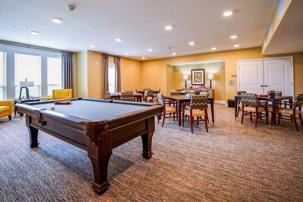 A cozy game room featuring a pool table, several tables and chairs, with warm lighting and carpeted flooring.