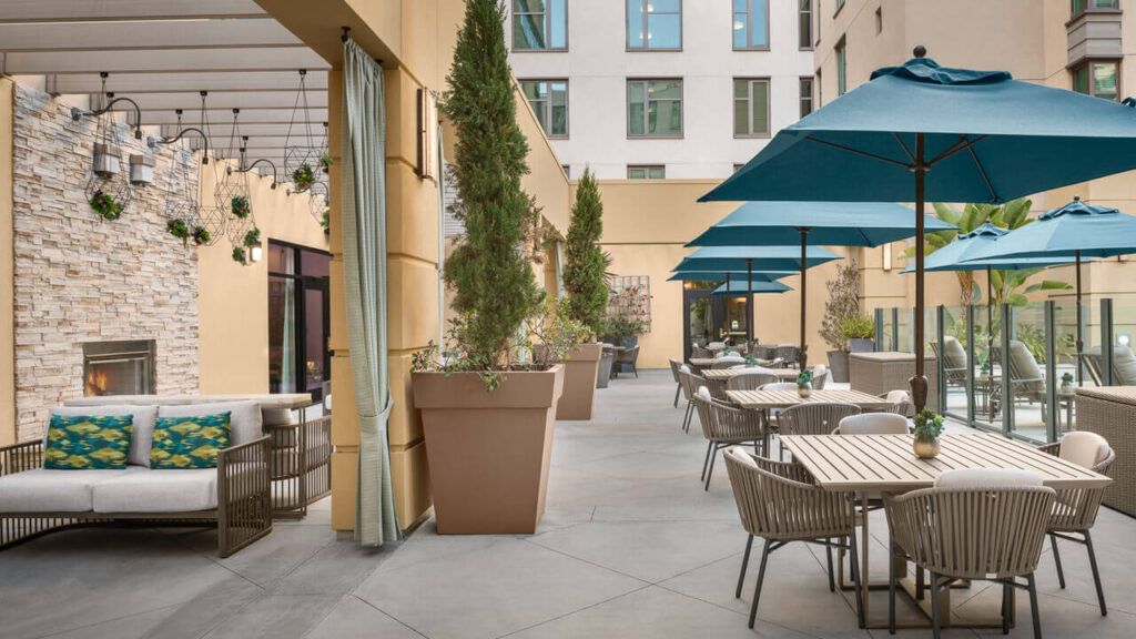 A stylish restaurant patio with tables, chairs, and umbrellas surrounded by tall potted plants in an urban setting.
