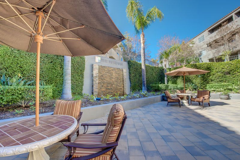 A sunny patio with outdoor furniture and umbrellas, surrounded by lush plants and palm trees.
