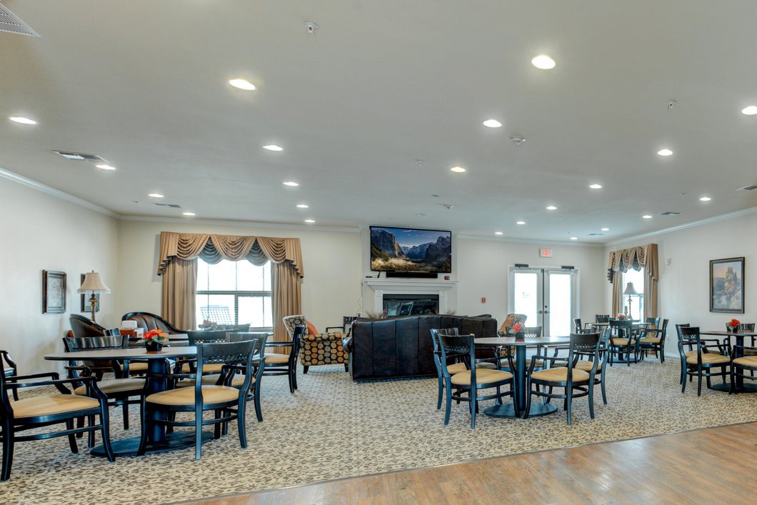 A spacious interior with a combined living and dining area, featuring several tables and chairs, a television mounted on the wall, and large windows providing natural light.