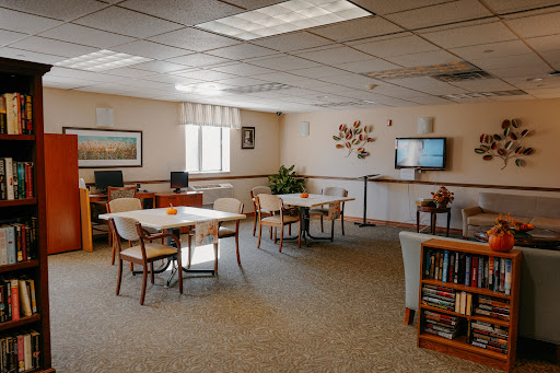 A spacious interior room featuring tables, chairs, a computer station, and a television, surrounded by bookshelves and natural light from the windows.